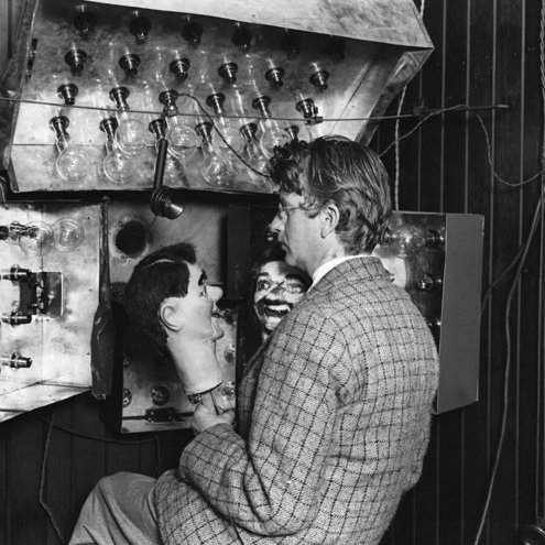 Black and white photograph of a person seated behind a microphone and a bank of light bulbs. They are holding a ventriloquist