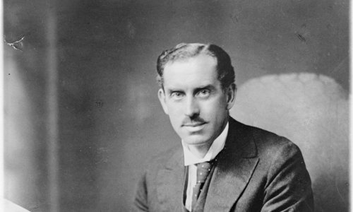 Black and white photograph of a person wearing a suit and tie seated at a table. They are looking into the camera and holding a large book in their hands, as if reading.