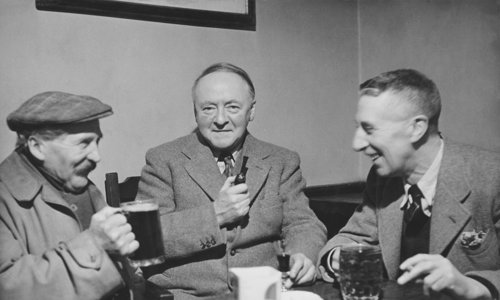 Black and white photo of three people seated at a table inside a public house. All are drinking and smiling, as if sharing a joke, and the person in the centre is looking at the camera and holding a pipe.