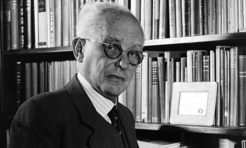 Black and white portrait photograph of a person standing in a library holding an open book. They are wearing a dark suit and tie and round glasses, and are looking directly at the camera.