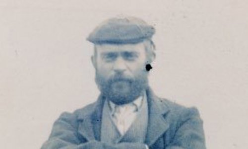 Black and white photograph taken in 1901 of Adam Christie. He is wearing a waistcoat and outer jacket, with a flat cap. His arms are folded and he is looking into the camera with a stern expression on his bearded face.