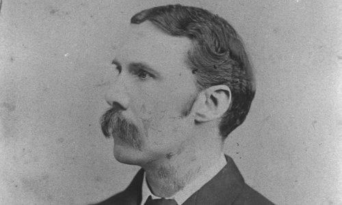 Black and white portrait photo of a person looking to the left, wearing a dark suit and tie. They have a large handlebar moustache.