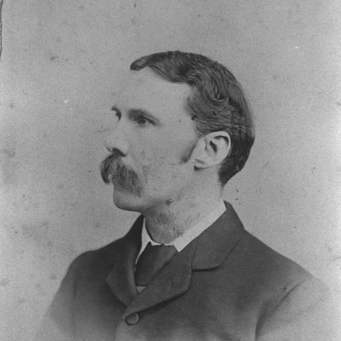 Black and white portrait photo of a person looking to the left, wearing a dark suit and tie. They have a large handlebar moustache.