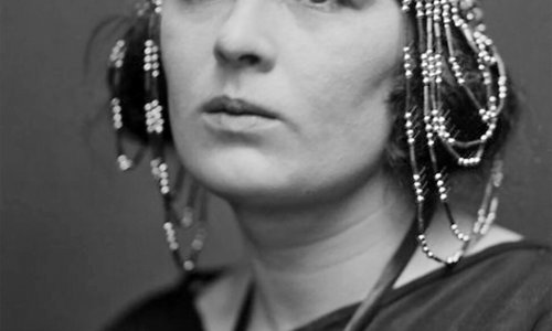 Black and white photograph of a person from the shoulders up. They are wearing a dark coloured top and an elaborate beaded headdress that runs across the forehead and falls down over both ears.