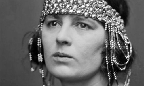Black and white photograph of a person from the shoulders up. They are wearing a dark coloured top and an elaborate beaded headdress that runs across the forehead and falls down over both ears.