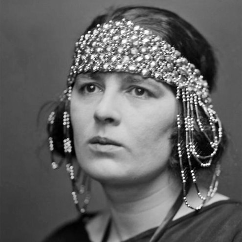 Black and white photograph of a person from the shoulders up. They are wearing a dark coloured top and an elaborate beaded headdress that runs across the forehead and falls down over both ears.
