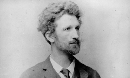 Black and white portrait photo of a person in a suit, jacket and tie. The person is looking off to the right and has unruly hair and a beard.