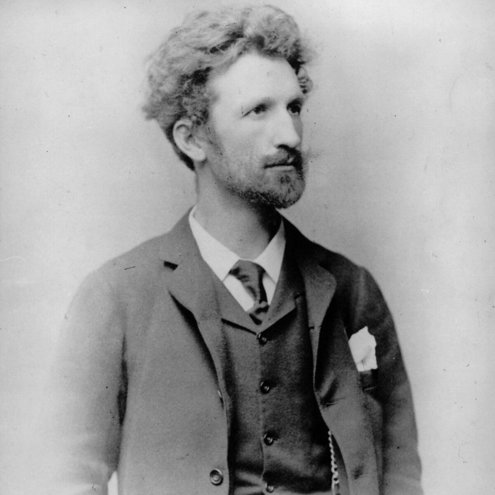 Black and white portrait photo of a person in a suit, jacket and tie. The person is looking off to the right and has unruly hair and a beard.
