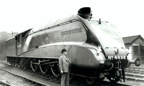 Black and white photograph of a person in a grey suit and dark trilby hat standing next to, and looking up at, a steam locomotive engine in a goods yard.