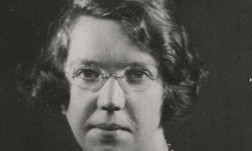 Black and white headshot portrait of a person wearing wire-rimmed glasses and a beaded necklace over a dark top.