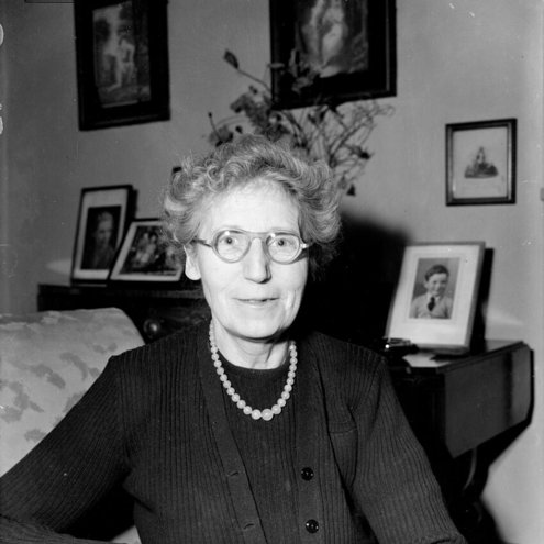 Black and white portrait photograph of a person sitting with their arms folded in an armchair. They are wearing glasses and a necklace, and behind them on a sideboard table are family photographs and a bunch of flowers in a vase.