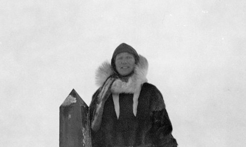 Black and white photo of a person standing with legs apart in deep snow, with one arm resting on a shoulder-high stone obelisk. They are wearing period-era Arctic clothing, possibly made from sealskin, and a big hat.