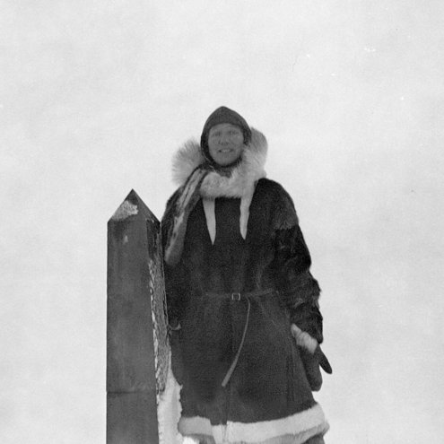 Black and white photo of a person standing with legs apart in deep snow, with one arm resting on a shoulder-high stone obelisk. They are wearing period-era Arctic clothing, possibly made from sealskin, and a big hat.