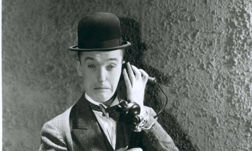 Black and white portrait photo of a person wearing a suit and bowler hat, pretending to talk on an old fashioned telephone. They have a sad or confused looking expression on their face.