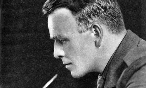 Black and white portrait photo of a person in military uniform, with side-parted slicked down hair. They are reading a book and smoking a pipe with a long stem.