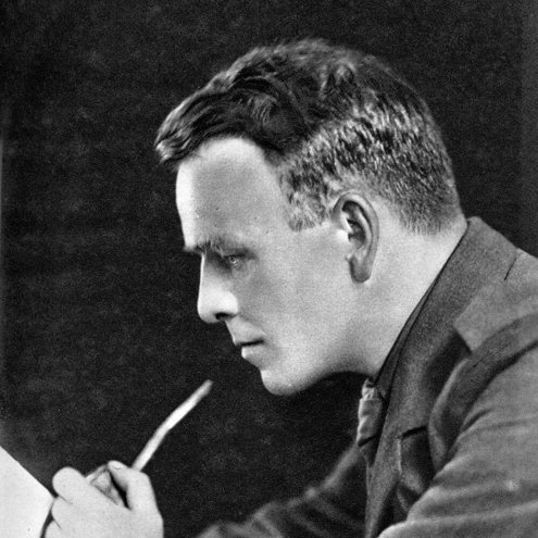 Black and white portrait photo of a person in military uniform, with side-parted slicked down hair. They are reading a book and smoking a pipe with a long stem.
