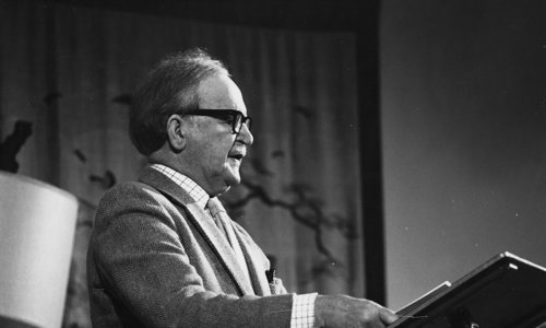 Black and white photo of a person giving a reading, standing at a lectern. They are wearing a jacket and tie and thick-rimmed black glasses. There is a book open in front of them.