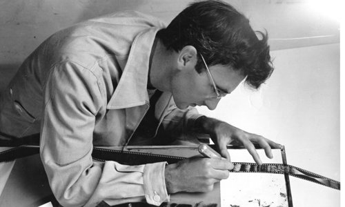 Black and white photo of a person in a shirt and glasses leaning over a table. They are marking a series of film negatives with a pen, as light shines upwards from underneath.