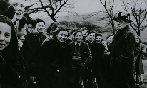 Black and white photo of a group of 1950s schoolchildren and a person supervising them. They are at a tree planting ceremony, and the uniformed children are laughing and smiling. The person is looking sternly at them, with their hands in their coat pockets.
