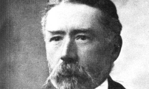 Black and white portrait photo of a person looking to the left of the camera. They are wearing a tweed jacket and have a beard and moustache.