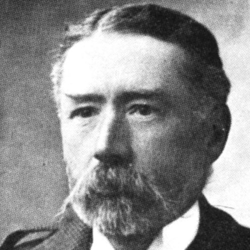 Black and white portrait photo of a person looking to the left of the camera. They are wearing a tweed jacket and have a beard and moustache.
