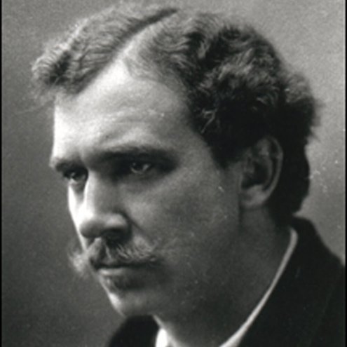 Black and white portrait photograph of a serious looking person wearing a suit and tie. They have a handlebar moustache and side parted hair, and they are staring off to the left.