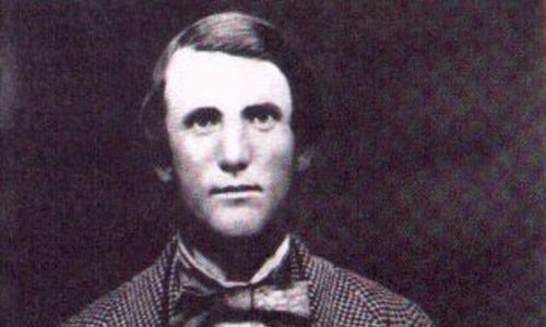 Black and white portrait photo of a person wearing a tweed suit and waistcoat. They also have a large dark bow tie and are looking at the camera with a relaxed expression, their right hand holding the lapel of their jacket.