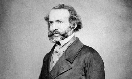Black and white portrait photo of a person wearing a fine suit and with elaborate mutton chop whiskers. They are standing with their right hand tucked inside their waistcoat.