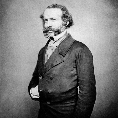 Black and white portrait photo of a person wearing a fine suit and with elaborate mutton chop whiskers. They are standing with their right hand tucked inside their waistcoat.