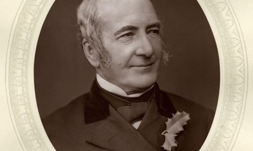 An early black and white portrait photo of a person looking off to the right. They are smiling, and wearing a dark suit, white shirt and black cravat, with two holly leaves pinned to their lapel. They have large grey sideburns reaching down to their cheeks.