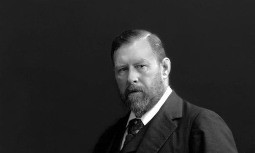 Black and white image of a seated man wearing a suit and tie. He has one hand on the arm of the chair and is looking at the camera.