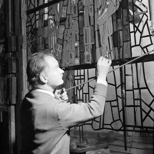 Black and white photograph of a person working on a very large stained glass window. They are carefully painting a section of the glass with a fine brush.