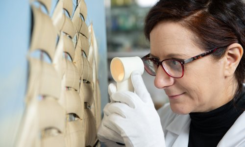 A person wearing glasses is looking closely at a painting of an old sailing ship with many sails. They are also wearing gloves and are looking intently at it through a small circular plastic instrument.