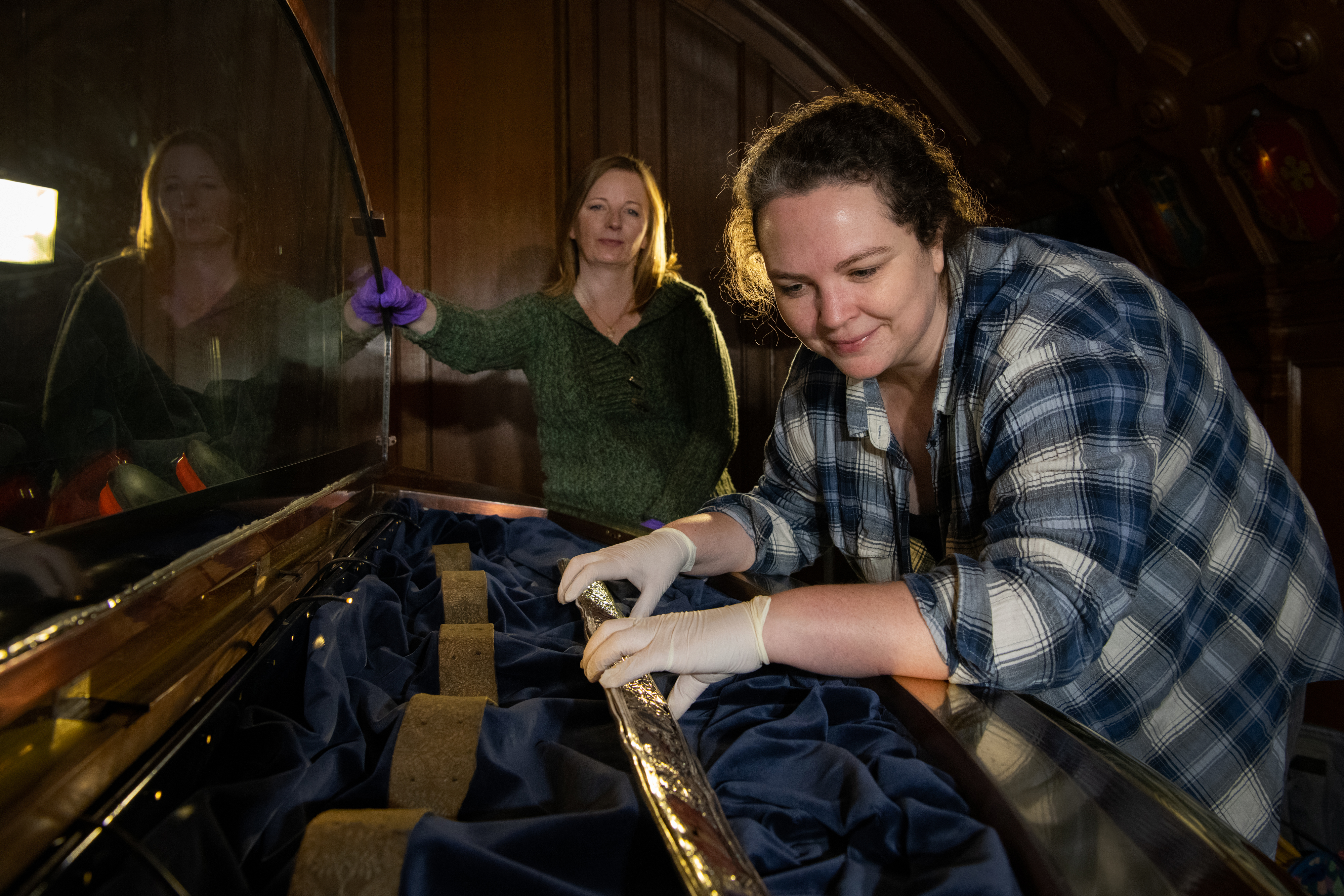 A conservator wearing protective gloves works inside a display case, carefully supporting the gold sceptre of Scotland on dark blue fabric while another conservator stands nearby.
