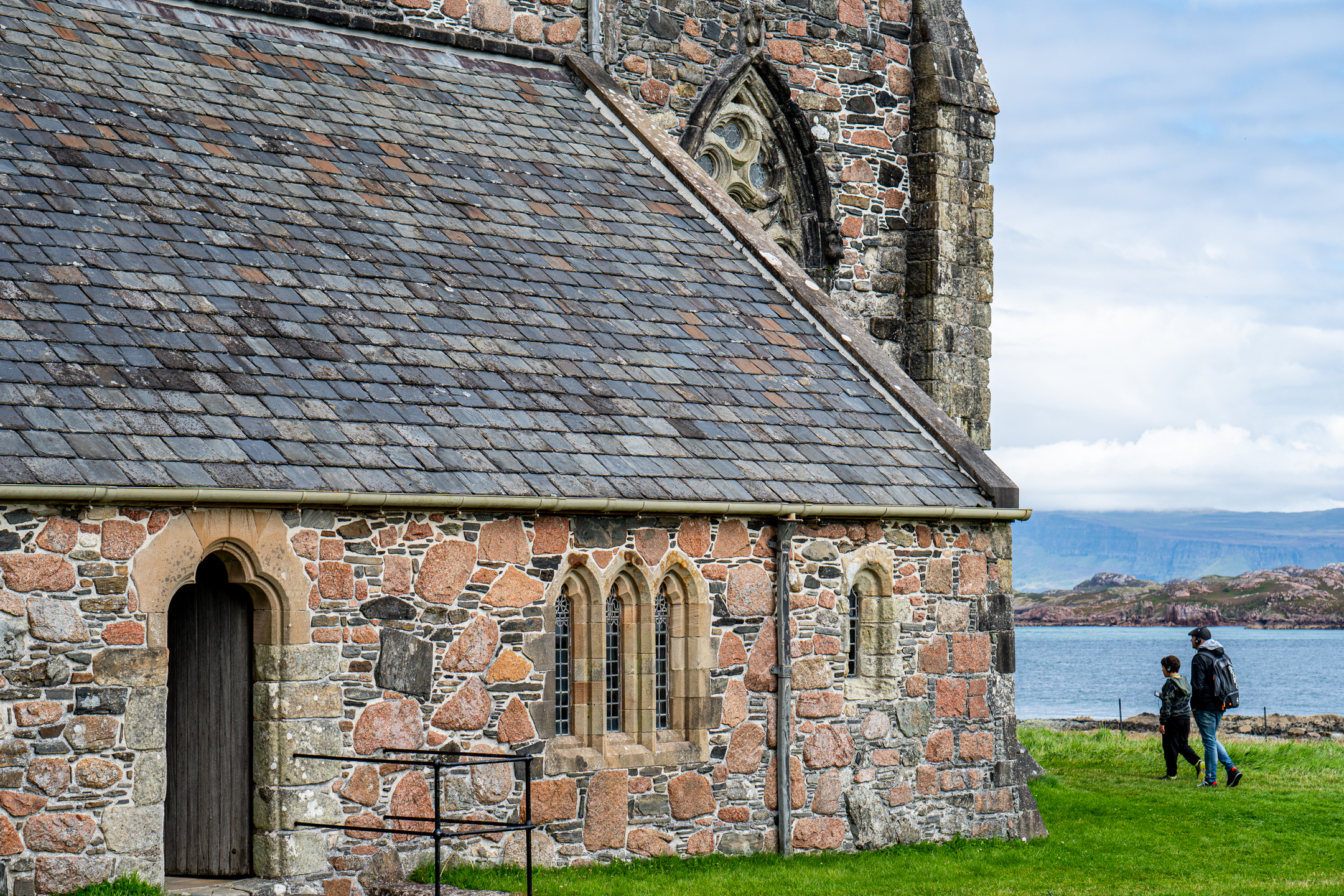 Part of a historic abbey with red, brown and yellow stones and a grey and red slate roof