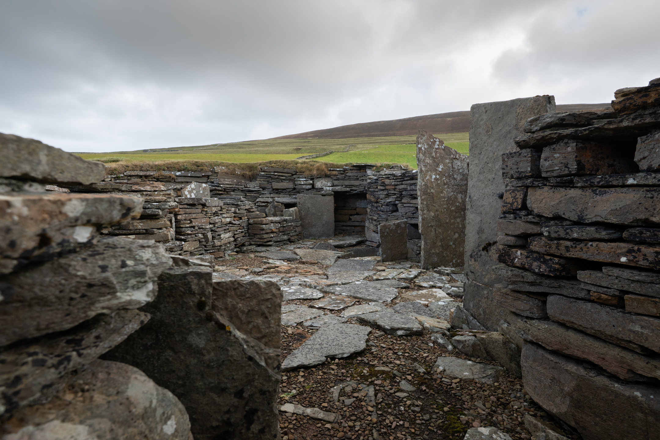 Interior view of Midhowe Broch showing low stone walls, upright slabs, and a paved floor with grassy hills visible through gaps in the ruins.