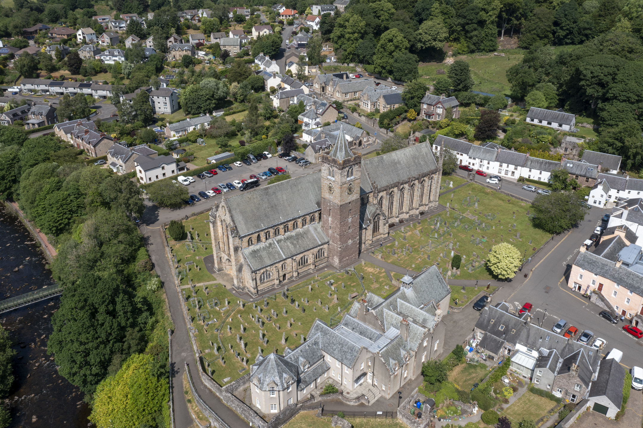 Aerial view of Dunblane Cathedral showing the long stone building with a central tower, surrounded by an expansive graveyard filled with rows of headstones, and bordered by nearby houses.