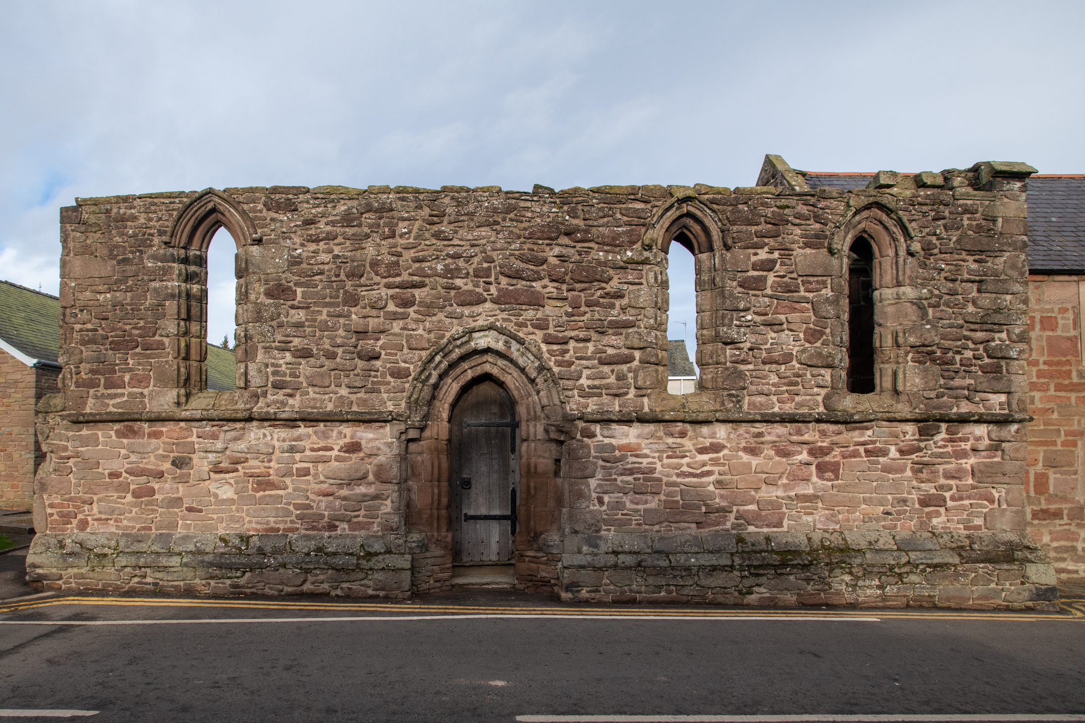 Closer view of the east gable of Maison Dieu Chapel showing the central pointed arch doorway and two tall lancet windows with chamfered stone surrounds. The stonework is irregular and weathered, with moss growth along the base.
