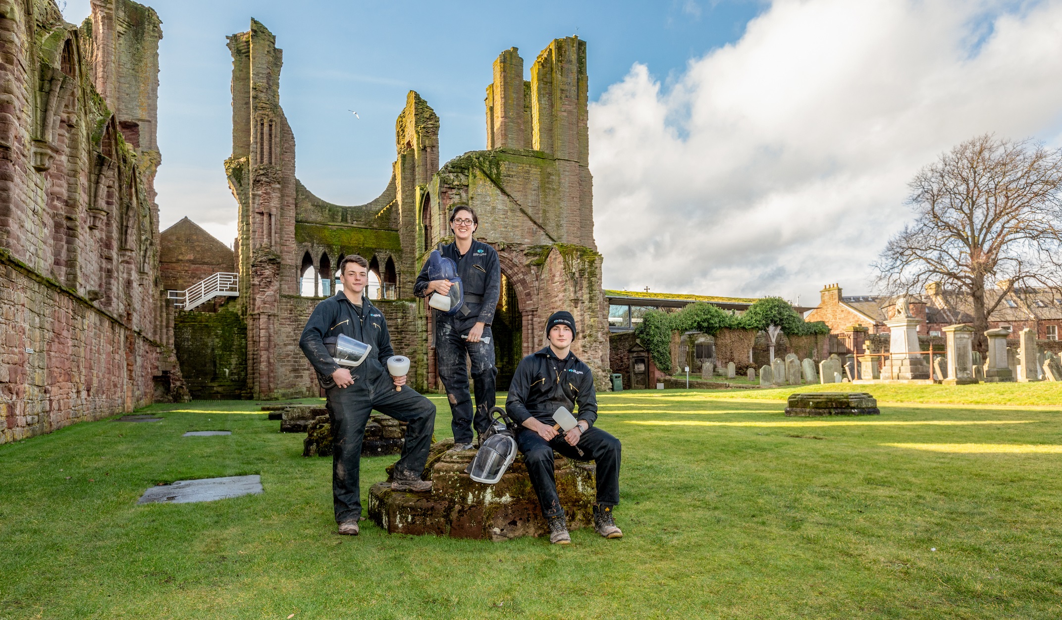 Portrait of apprentice stonemasons at Arbroath Abbey. Liam Macaulay, stonemason, Carolann Neilson, and apprentice Luke Maher pose with conservation tools while standing and sitting on historic stonework within the abbey ruins. The red sandstone walls and arches of Arbroath Abbey rise behind them, with gravestones, a grassy courtyard, and a bright sky with scattered clouds completing the scene.