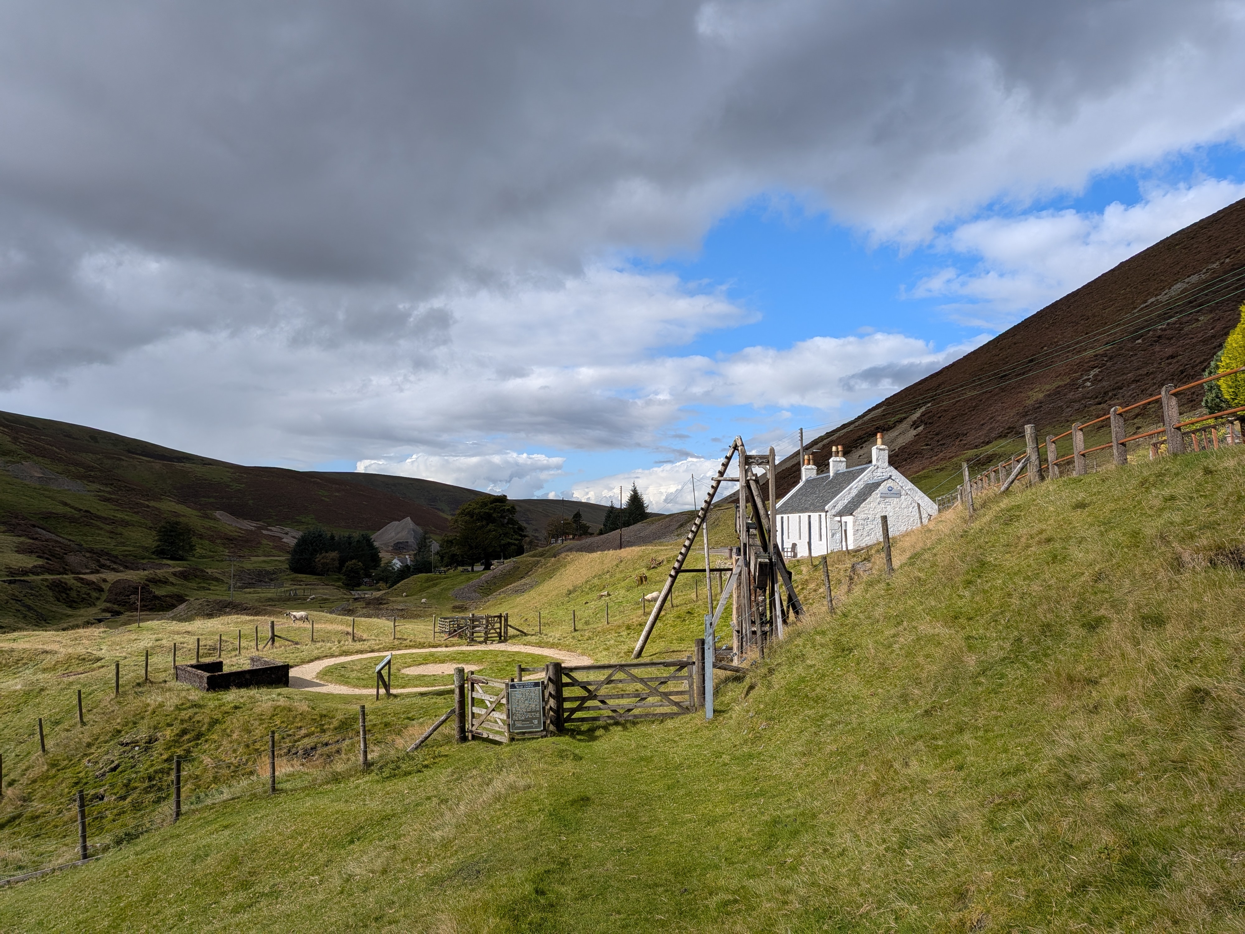 A panoramic photo of a desolate glen. In the foreground there is a wooden pump, a relic from the mining industry which once thrived in the glen. Beside the beam engine there is a small stone cottage, 