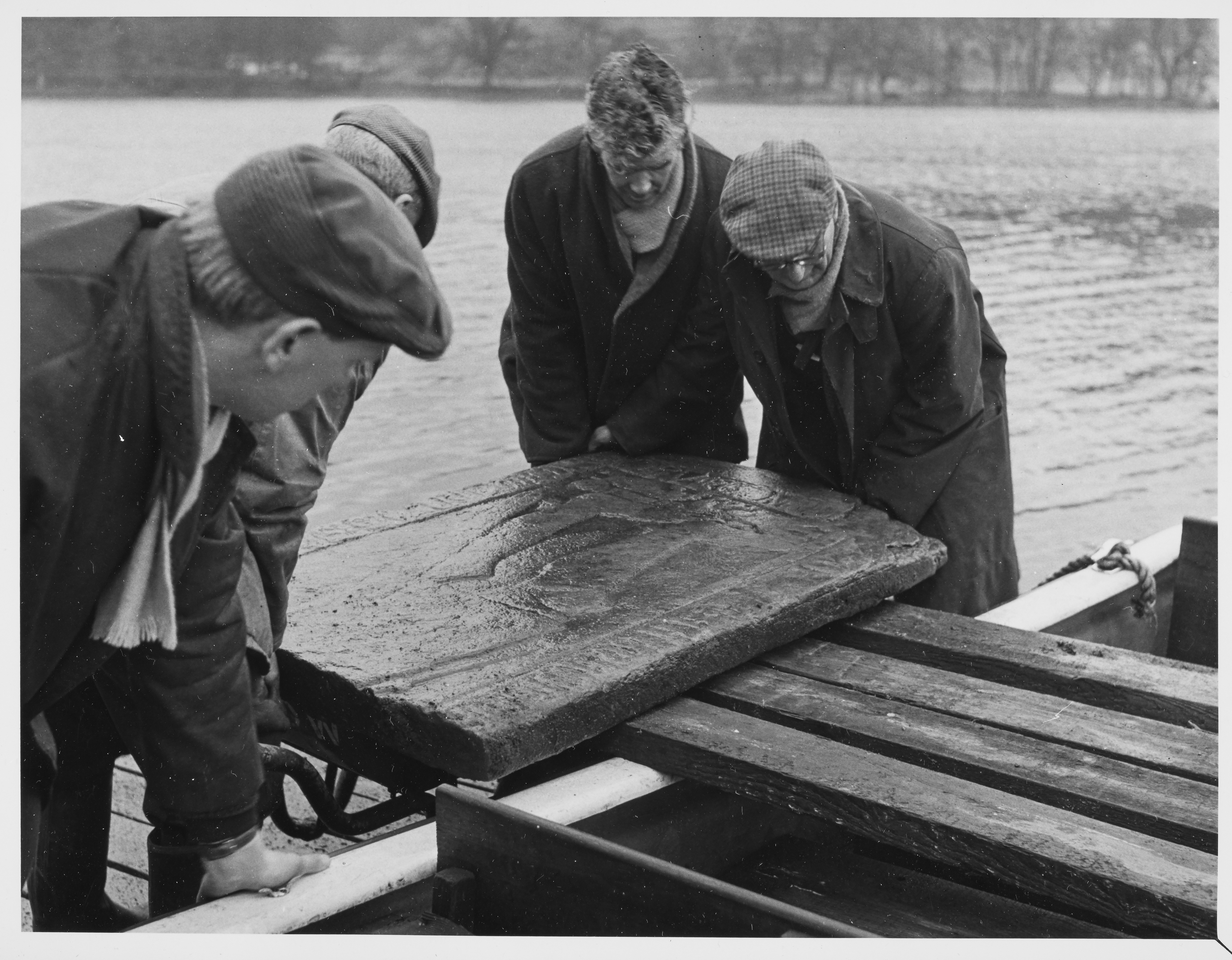 Four men move a carved stone slab on or off a boat using wooden planks for support