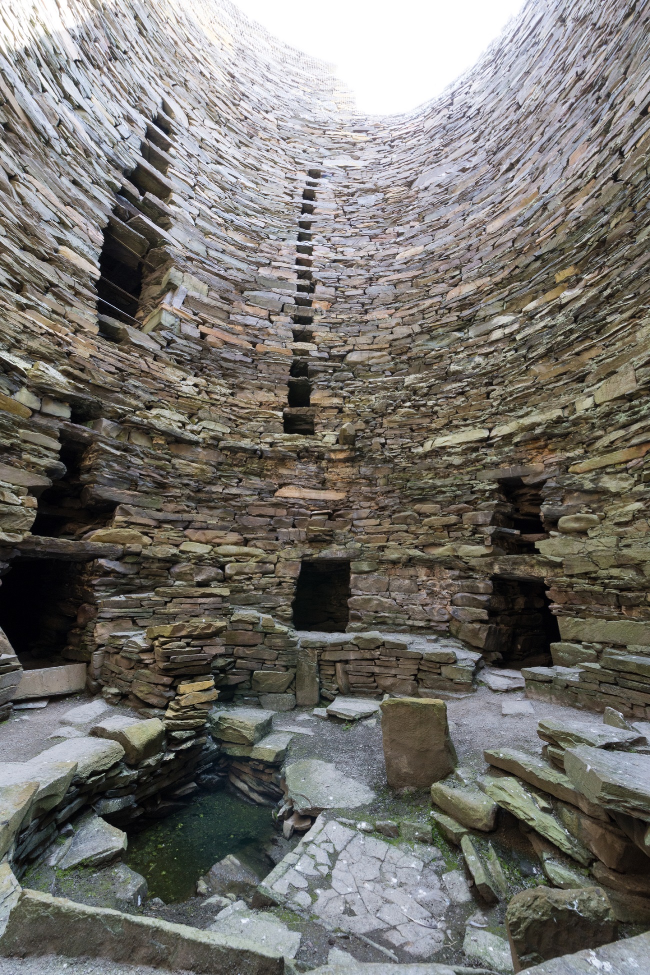 Interior base of Mousa Broch, with layered drystone walls, small chambers built into the structure, and debris scattered on the ground.