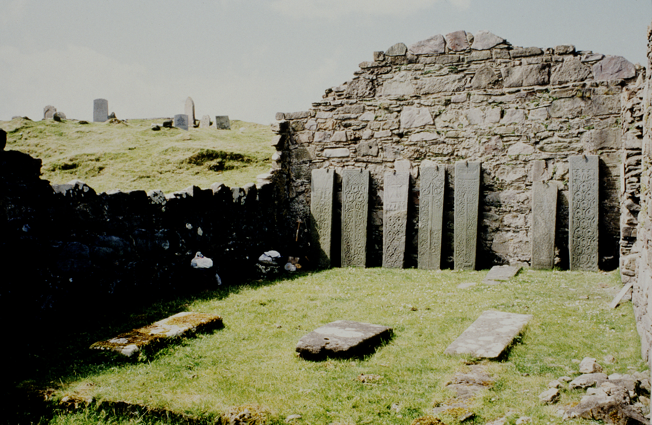 Interior of the chapel ruins showing six upright carved grave slabs lined against a stone wall, with grass and scattered stones on the floor.