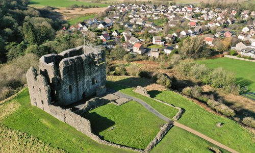 A ruined castle on a hill, with a modern housing estate in the background. The castle is a tall rectangular grey building without a roof surrounded by a low wall.