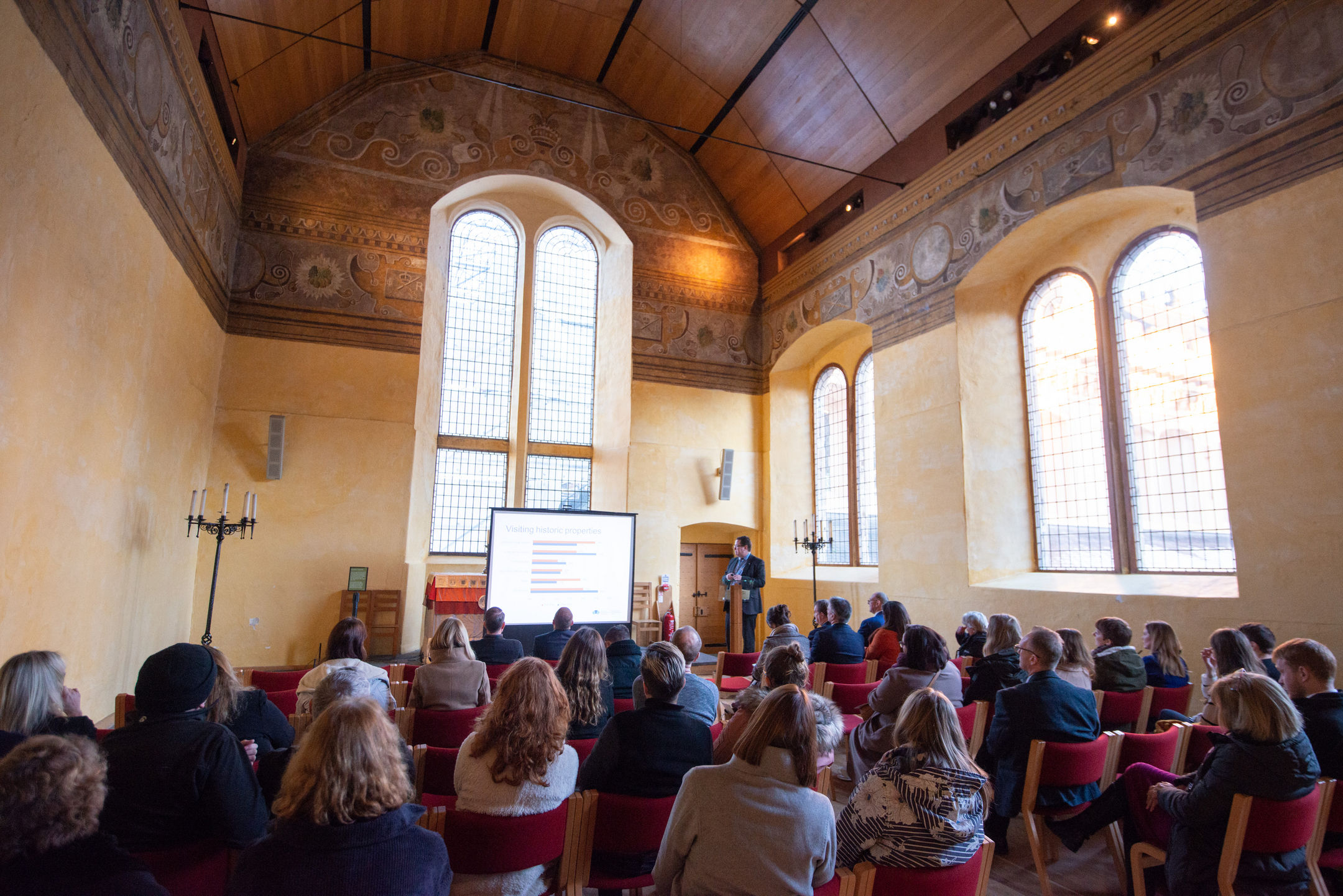 A large group of people all sit on red chairs facing a screen and presenter at the front of the room. The room has a high wooden vaulted ceiling and painted plaster.