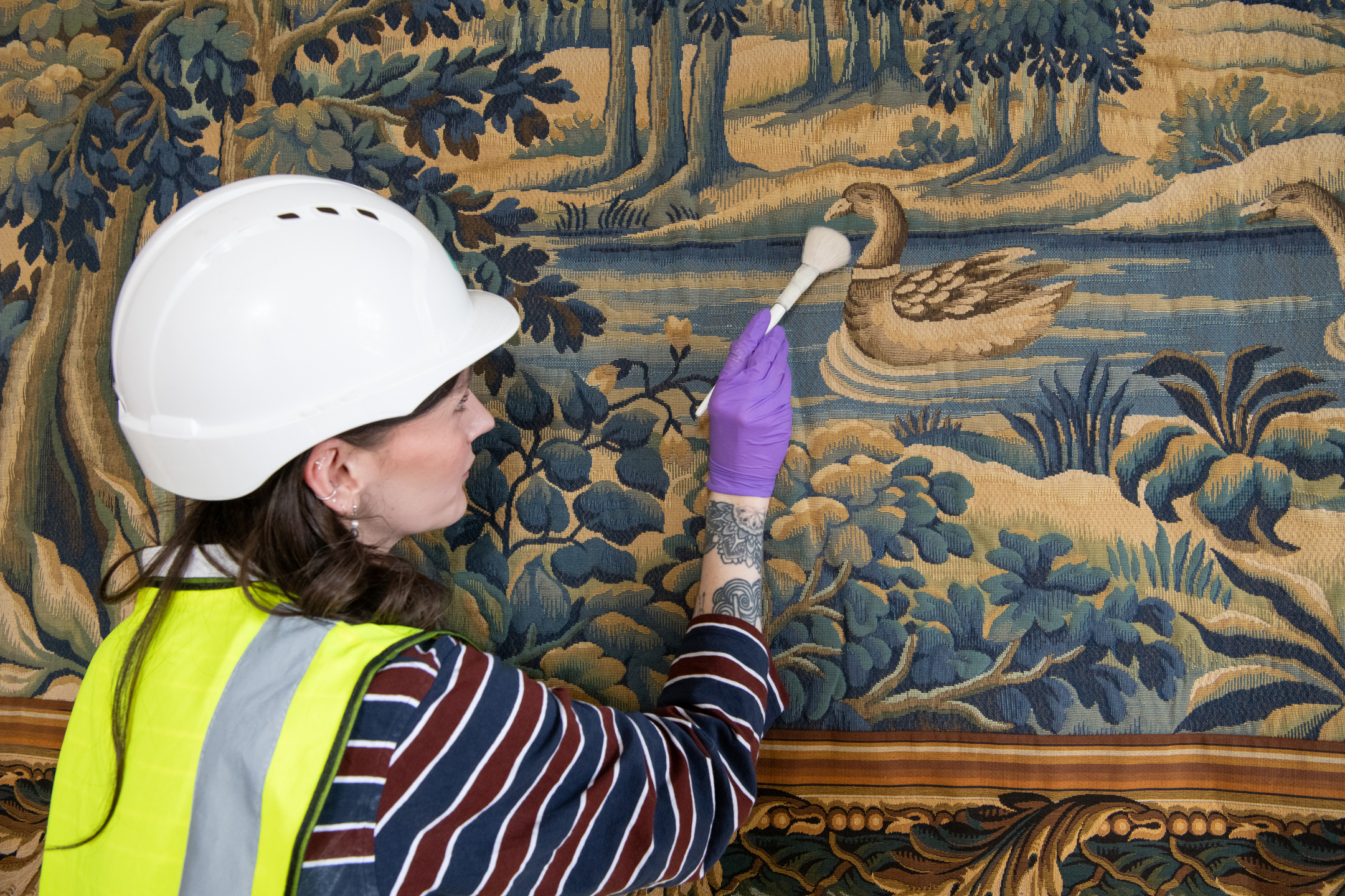 A member of the Historic Environment Scotland Collections team uses a small brush to clean a large tapestry on which there is a depiction of a duck on a river. 