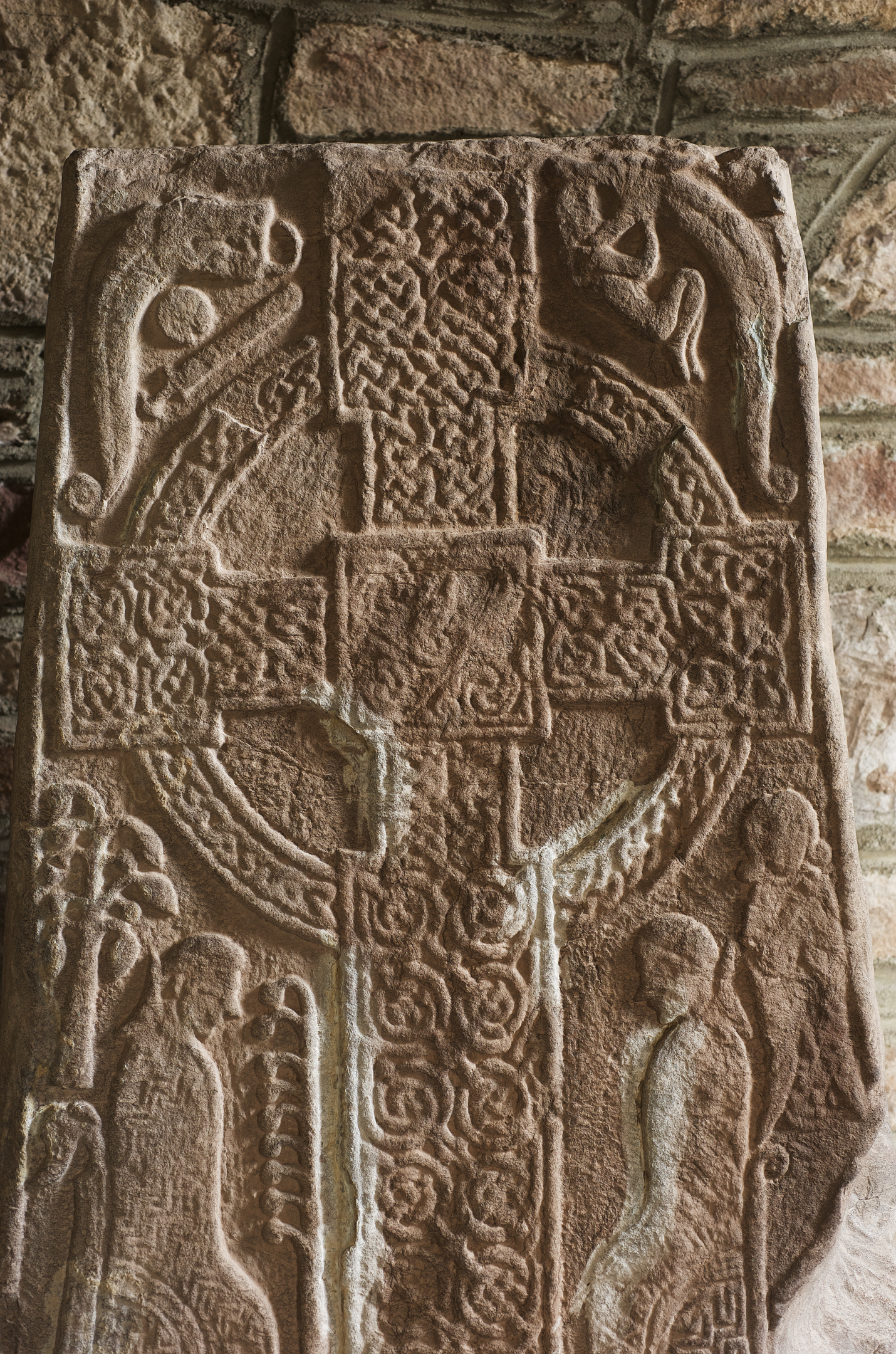 Close-up of the upper portion of a carved stone cross-slab featuring a Celtic cross design with interlaced knotwork and stylized animal motifs. The carvings are symmetrical and deeply cut, with a circular ring around the cross. The stone is set against a background of rough masonry