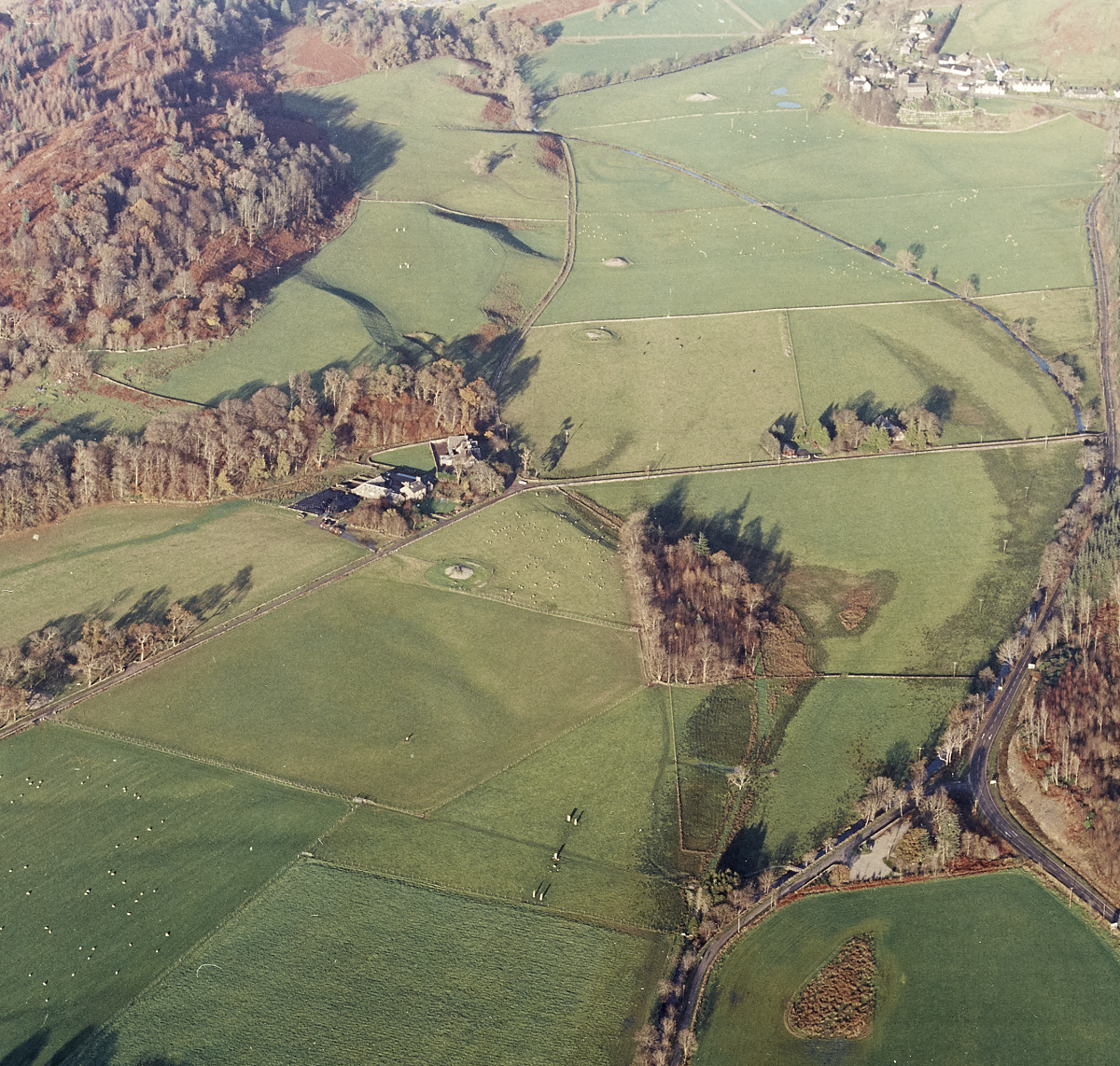 A wide aerial view of Kilmartin Glen showing part of the linear cemetery: Glebe Cairn at the top, followed by Nether Largie North Cairn, Nether Largie Mid Cairn, and Nether Largie South Cairn near the bottom. To the right of the image, the Nether Largie Standing Stones are visible in a field. The cairns and stones sit within a patchwork of green fields, woodland, and farm tracks.