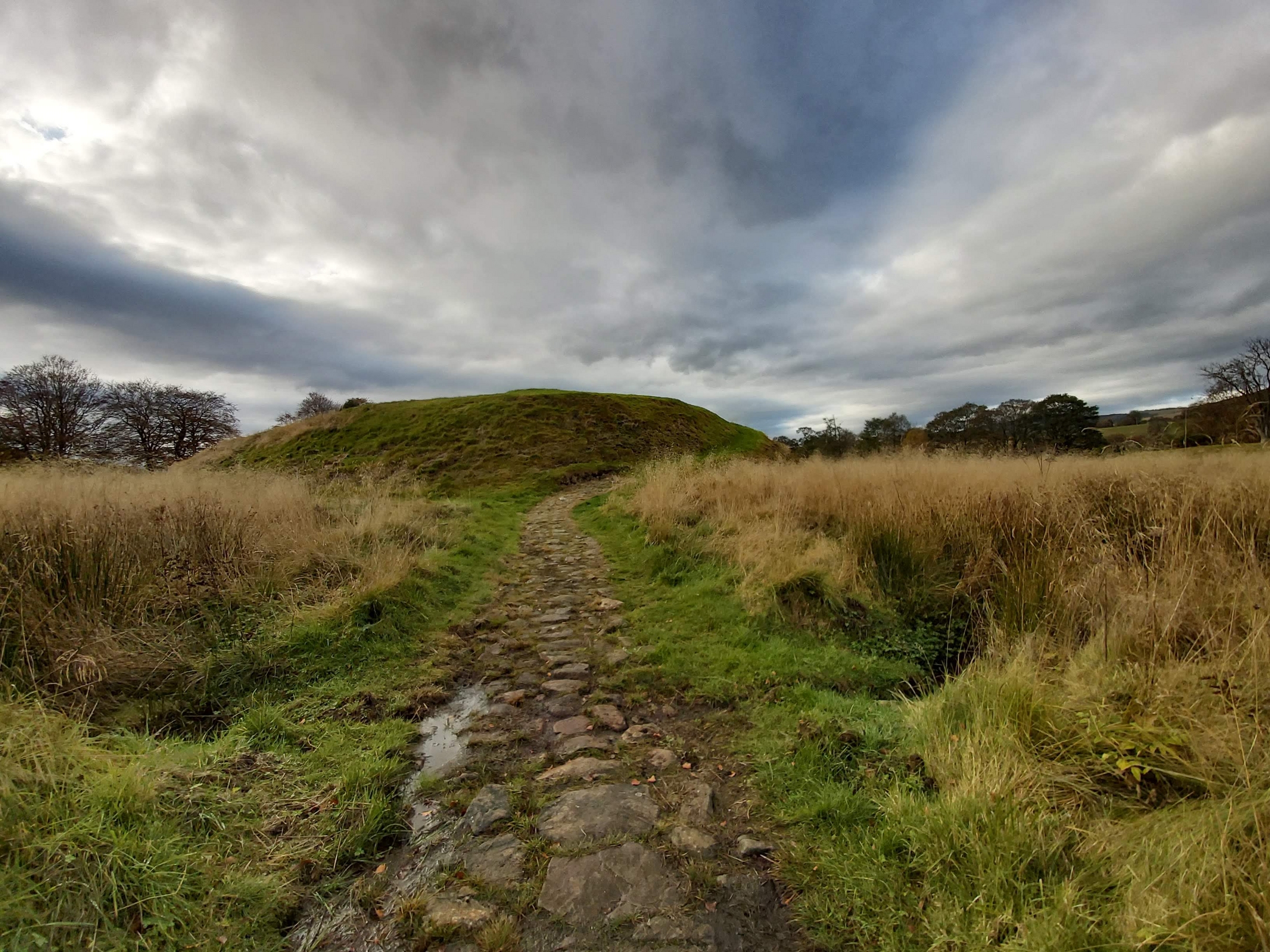 A stone path leading through grassy terrain toward the Peel Ring of Lumphanan under a dramatic cloudy sky.