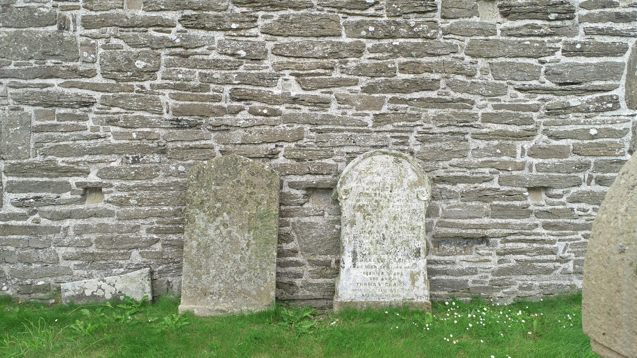 Two weathered gravestones positioned against a rough‑stone exterior wall of the church.